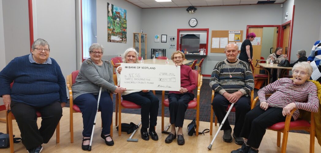 group of people sitting in a semi circle, three ladies holding a large cheque