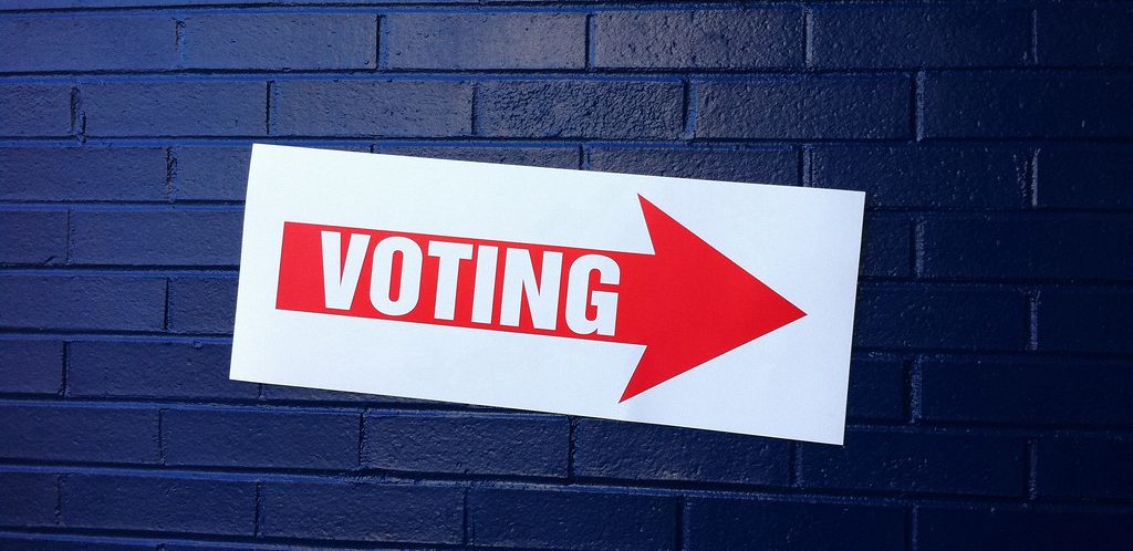 White poster with red arrow pointing to the right with the word voting, against a dark blue brick wall