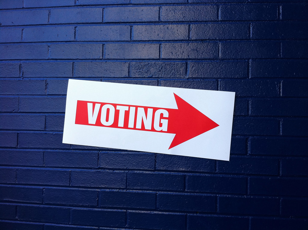 White poster with red arrow pointing to the right with the word voting, against a dark blue brick wall