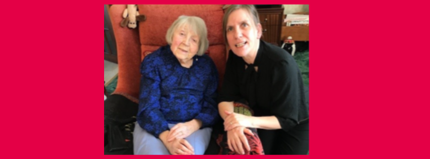 Aileen, and older woman in blue jumper, sits in a chair and Gill crouches next to her, wearing a black top