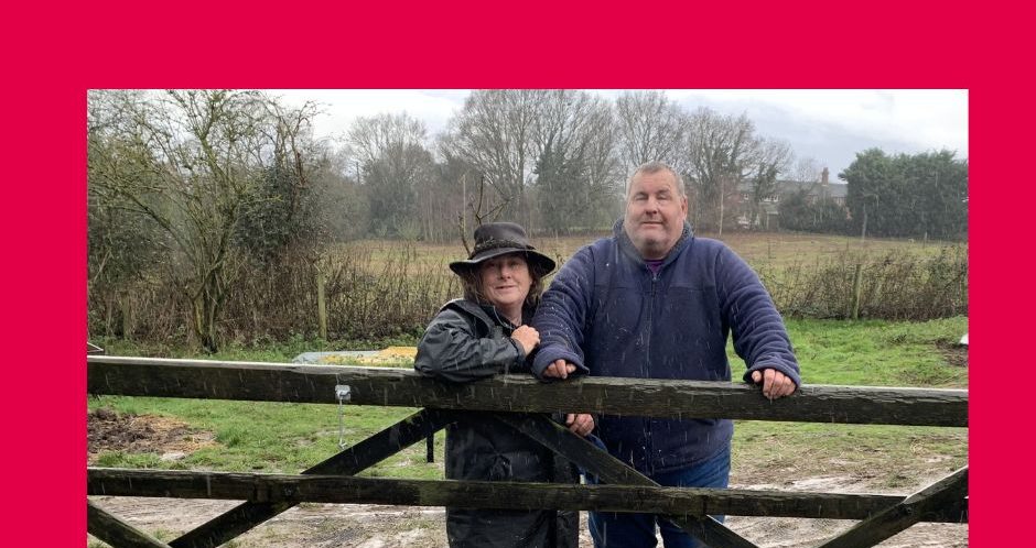 Mike and NESS who run Inclusive Farms stand behind a five bar farm gate in a muddy field